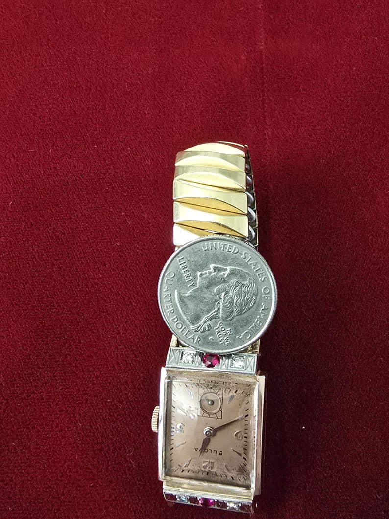 May include: Vintage Bulova watch with a rectangular face, gold-tone band, and a quarter coin placed on top. The watch face has a light-colored background with black hands and numerals. Red gemstones are set on the watch.