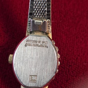 K&ouml;nnte beinhalten: Nahaufnahme einer Vintage-Armbanduhr mit einem silberfarbenen Metallmaschenband. Das Zifferblatt der Uhr ist silberfarben mit einer goldfarbenen Krone. Auf der R&uuml;ckseite der Uhr steht "STAINLESS ST. BACK" und "BASE METAL BEZEL".