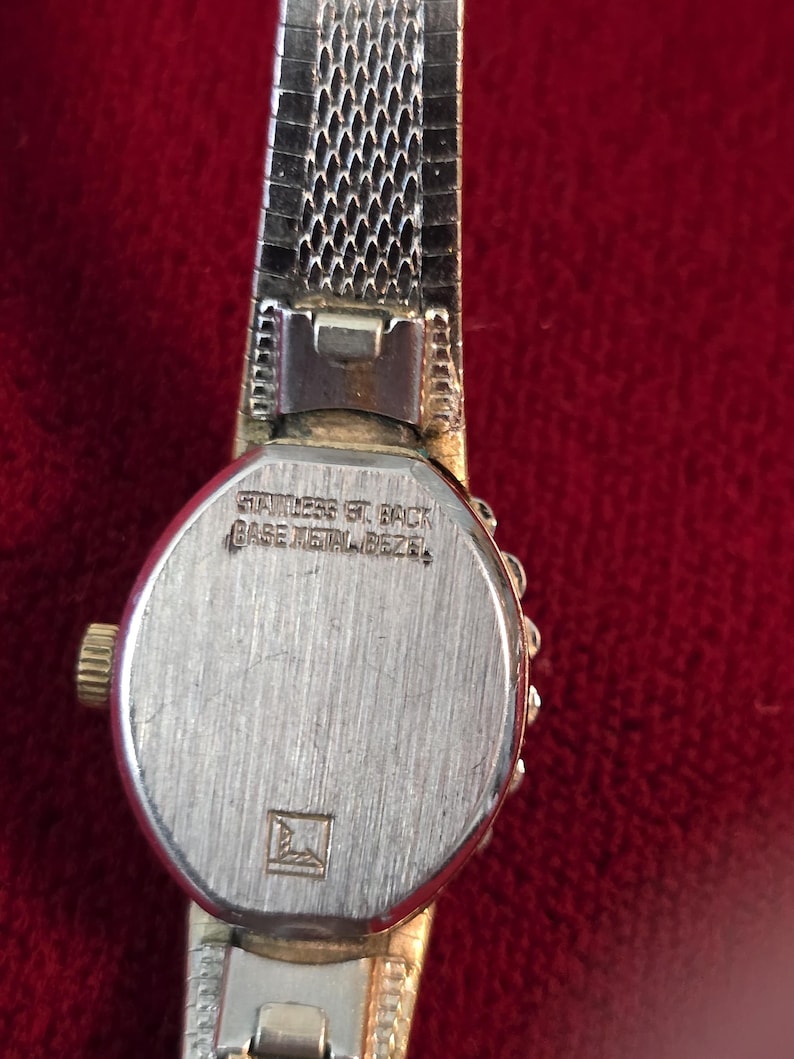 K&ouml;nnte beinhalten: Nahaufnahme einer Vintage-Armbanduhr mit einem silberfarbenen Metallmaschenband. Das Zifferblatt der Uhr ist silberfarben mit einer goldfarbenen Krone. Auf der R&uuml;ckseite der Uhr steht "STAINLESS ST. BACK" und "BASE METAL BEZEL".