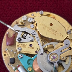 Puede incluir: Primer plano de un movimiento de reloj vintage con engranajes, tornillos y un volante dorados. El mecanismo del reloj tiene un componente de color turquesa y el texto "THIRTEEN 13 JEWELS SWISS". El reloj est&aacute; etiquetado como "Sears Roebuck Co Electronic".