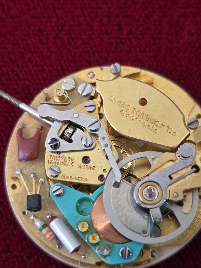 Puede incluir: Primer plano de un movimiento de reloj vintage con engranajes, tornillos y un volante dorados. El mecanismo del reloj tiene un componente de color turquesa y el texto "THIRTEEN 13 JEWELS SWISS". El reloj est&aacute; etiquetado como "Sears Roebuck Co Electronic".