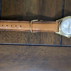 May include: A vintage gold-toned wristwatch with a light-colored face and a brown leather band. The watch has a classic design with a round face and a buckle closure. The watch band is approximately 0.75 inches wide.