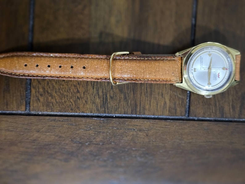May include: A vintage gold-toned wristwatch with a light-colored face and a brown leather band. The watch has a classic design with a round face and a buckle closure. The watch band is approximately 0.75 inches wide.
