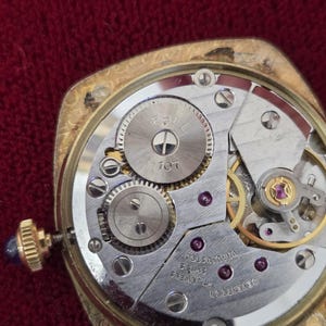 May include: Close-up of a vintage gold-toned watch mechanism. The watch face is open, revealing the intricate internal gears, screws, and jewels. The watch has a gold crown with a blue stone. The watch is marked "EPEL" and "17 JEWELS SWISS".