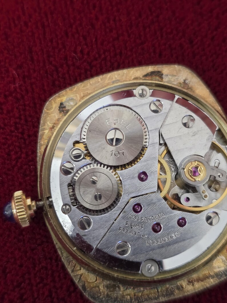 May include: Close-up of a vintage gold-toned watch mechanism. The watch face is open, revealing the intricate internal gears, screws, and jewels. The watch has a gold crown with a blue stone. The watch is marked "EPEL" and "17 JEWELS SWISS".