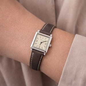 May include: A Bulova watch with a square, silver-toned face and a brown leather band. The watch face is cream-colored with black numerals and hands. The band is dark brown with white stitching.