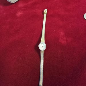 K&ouml;nnte beinhalten: Eine goldfarbene Armbanduhr mit einem filigranen Armband im Netzstil. Das Zifferblatt der Uhr ist rund, umgeben von einem Kranz aus kleinen, klaren Steinen. Die Uhr liegt auf rotem Untergrund.