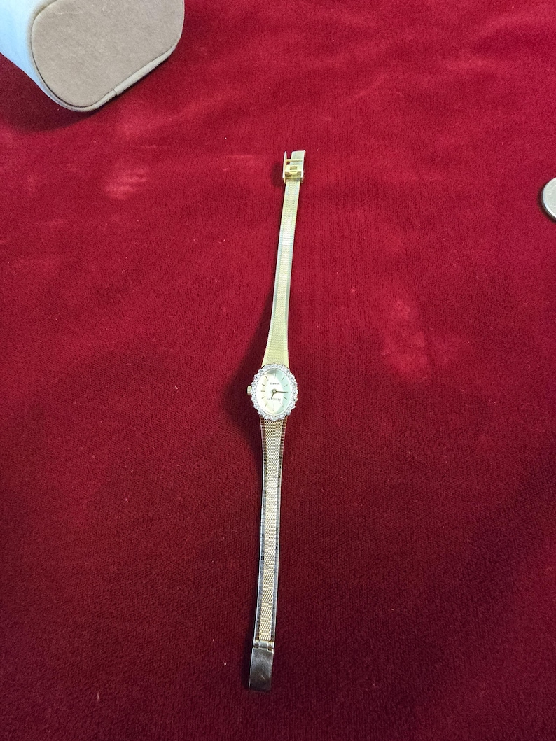 K&ouml;nnte beinhalten: Eine goldfarbene Armbanduhr mit einem filigranen Armband im Netzstil. Das Zifferblatt der Uhr ist rund, umgeben von einem Kranz aus kleinen, klaren Steinen. Die Uhr liegt auf rotem Untergrund.