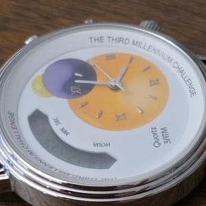 Op de afbeelding: Een zilverkleurig analoog en digitaal horloge met een witte wijzerplaat met de tekst THE THIRD MILLENNIUM CHALLENGE. De wijzerplaat heeft een geel-naar-oranje verloop in het midden met Romeinse cijfers en een paarse en gele planeetgrafiek.