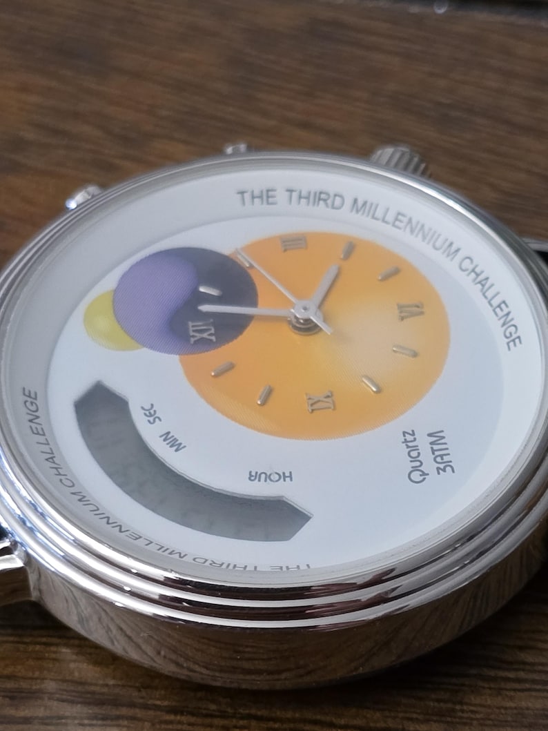 Op de afbeelding: Een zilverkleurig analoog en digitaal horloge met een witte wijzerplaat met de tekst THE THIRD MILLENNIUM CHALLENGE. De wijzerplaat heeft een geel-naar-oranje verloop in het midden met Romeinse cijfers en een paarse en gele planeetgrafiek.