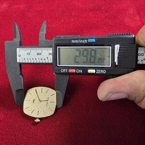 May include: A gold-toned watch face with a square shape is measured with a digital caliper. The watch face is approximately 1.17 inches in length. The watch has a small crown and a simple, elegant design.