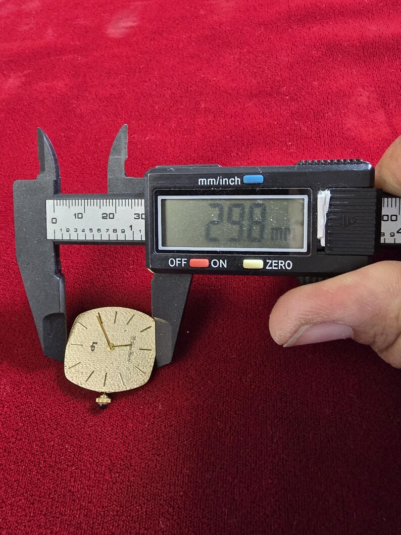 May include: A gold-toned watch face with a square shape is measured with a digital caliper. The watch face is approximately 1.17 inches in length. The watch has a small crown and a simple, elegant design.