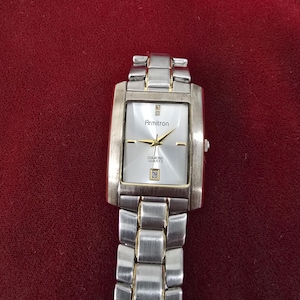 May include: Silver-toned Armitron Diamond Quartz watch with a rectangular face and a silver link band. The watch face is white with gold-colored hands and markers. The date is displayed at the 3 o'clock position. The watch is on a red background.