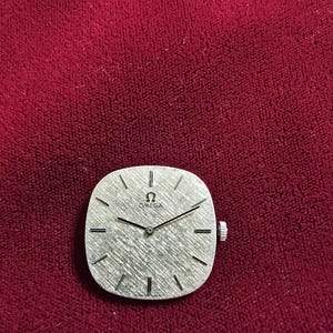 Pu&ograve; includere: Un orologio da polso Omega vintage con quadrante argentato strutturato e lancette nere. L'orologio ha una forma quadrata con bordi arrotondati e una corona argentata. Lo sfondo &egrave; un tessuto rosso scuro.