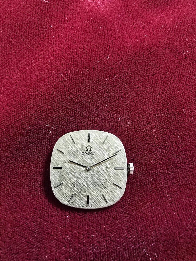 Pu&ograve; includere: Un orologio da polso Omega vintage con quadrante argentato strutturato e lancette nere. L'orologio ha una forma quadrata con bordi arrotondati e una corona argentata. Lo sfondo &egrave; un tessuto rosso scuro.