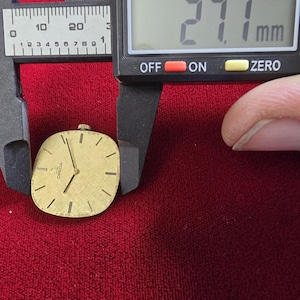 Pu&ograve; includere: Un quadrante di orologio Omega dorato con lancette e indici neri, di circa 27,1 mm. Il quadrante &egrave; su uno sfondo rosso, con un calibro digitale che mostra la misura in millimetri.