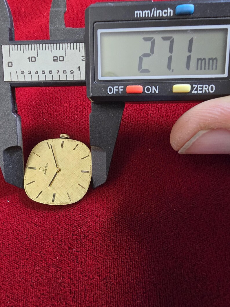 Pu&ograve; includere: Un quadrante di orologio Omega dorato con lancette e indici neri, di circa 27,1 mm. Il quadrante &egrave; su uno sfondo rosso, con un calibro digitale che mostra la misura in millimetri.