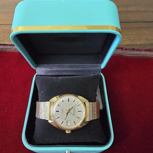 May include: A gold-toned wristwatch with a mesh band rests on a black pillow inside a turquoise box with a gold trim. The watch has a light-colored face with black markers and a date window. The box is open, revealing the watch and its presentation.