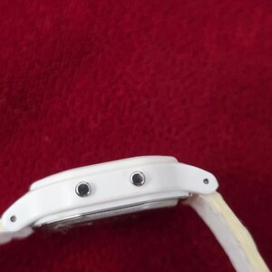 May include: A white digital watch with a light-colored band. The watch face has two black buttons and a silver-colored backing. The watch is set against a red background.