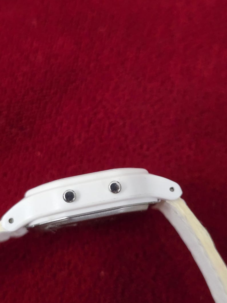 May include: A white digital watch with a light-colored band. The watch face has two black buttons and a silver-colored backing. The watch is set against a red background.