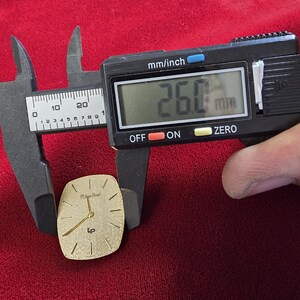 May include: A gold-toned watch face with a rectangular shape, featuring gold hands and hour markers. The watch face is measured with a digital caliper, showing a width of 26.0 mm, or approximately 1.02 inches. The background is a deep red fabric.