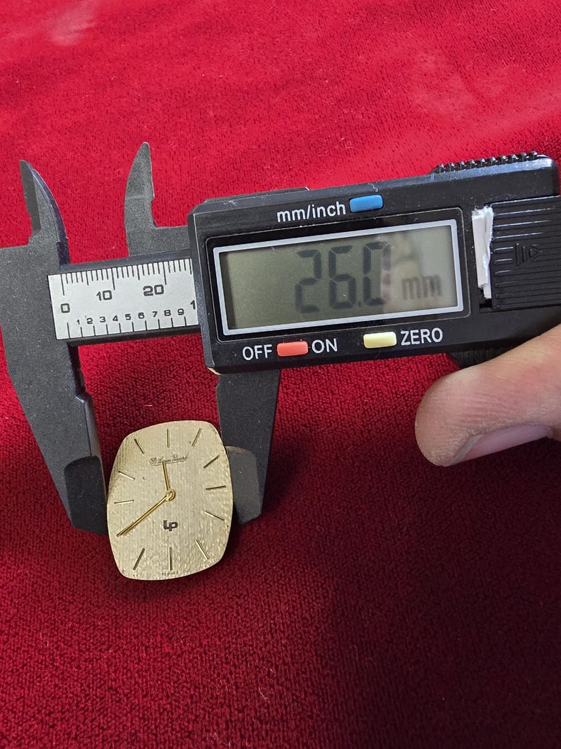 May include: A gold-toned watch face with a rectangular shape, featuring gold hands and hour markers. The watch face is measured with a digital caliper, showing a width of 26.0 mm, or approximately 1.02 inches. The background is a deep red fabric.