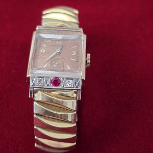 May include: A vintage Bulova wristwatch with a rectangular gold-tone face and a gold band. The watch face is a light tan color with gold hands and hour markers. The band features a decorative section with a ruby and diamonds.