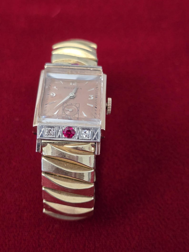May include: A vintage Bulova wristwatch with a rectangular gold-tone face and a gold band. The watch face is a light tan color with gold hands and hour markers. The band features a decorative section with a ruby and diamonds.