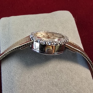 K&ouml;nnte beinhalten: Eine goldfarbene Armbanduhr mit rundem Zifferblatt und einer L&uuml;nette, die mit kleinen, klaren Edelsteinen besetzt ist. Die Uhr hat ein goldenes Mesh-Armband und eine kleine, goldene Krone. Die Uhr liegt auf einer grauen Oberfl&auml;che vor rotem Hintergrund.