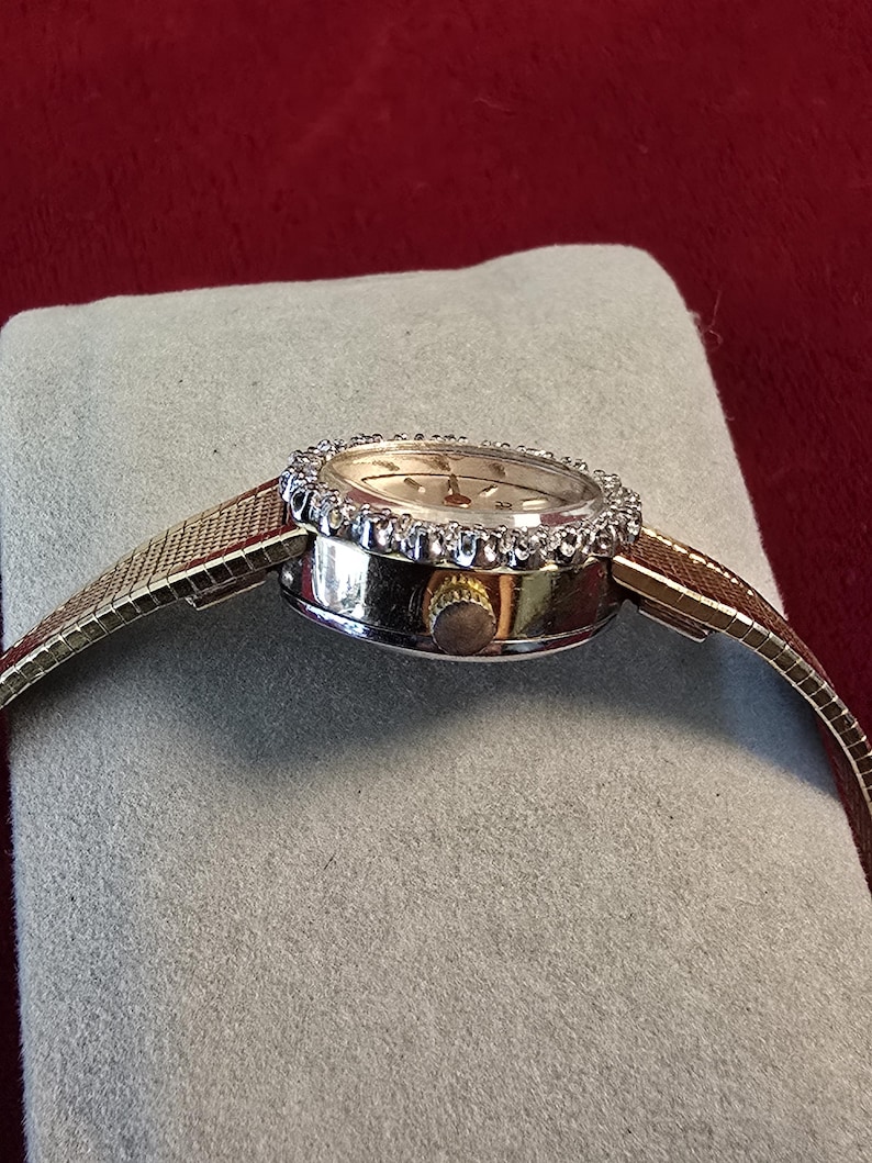 K&ouml;nnte beinhalten: Eine goldfarbene Armbanduhr mit rundem Zifferblatt und einer L&uuml;nette, die mit kleinen, klaren Edelsteinen besetzt ist. Die Uhr hat ein goldenes Mesh-Armband und eine kleine, goldene Krone. Die Uhr liegt auf einer grauen Oberfl&auml;che vor rotem Hintergrund.