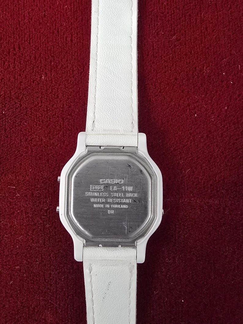 May include: A white Casio LA-11W digital watch with a white leather strap. The watch has a stainless steel back and is water resistant. The watch is made in Thailand.