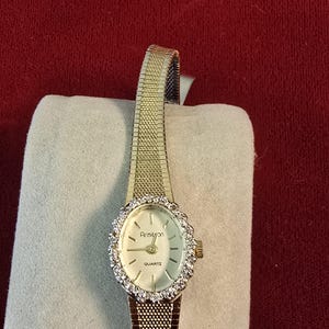 K&ouml;nnte beinhalten: Eine goldfarbene Armitron-Quarzuhr mit einem Mesh-Armband. Das ovale Zifferblatt ist von einem Ring aus klaren Steinen umgeben. Die Zeiger der Uhr sind schwarz, und das Zifferblatt hat eine hellcremefarbene Farbe.