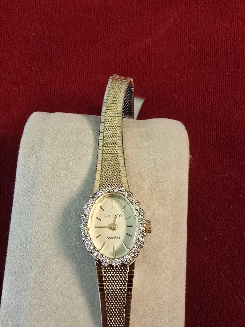 K&ouml;nnte beinhalten: Eine goldfarbene Armitron-Quarzuhr mit einem Mesh-Armband. Das ovale Zifferblatt ist von einem Ring aus klaren Steinen umgeben. Die Zeiger der Uhr sind schwarz, und das Zifferblatt hat eine hellcremefarbene Farbe.