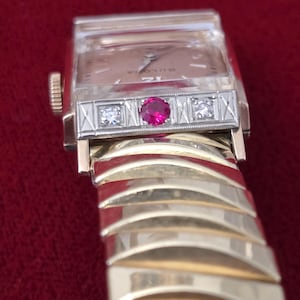 May include: A vintage Bulova wristwatch with a gold-tone band and case. The watch face is rectangular with a light-colored dial. The bezel features a central ruby and two small diamonds. The band is a series of curved gold-tone links.