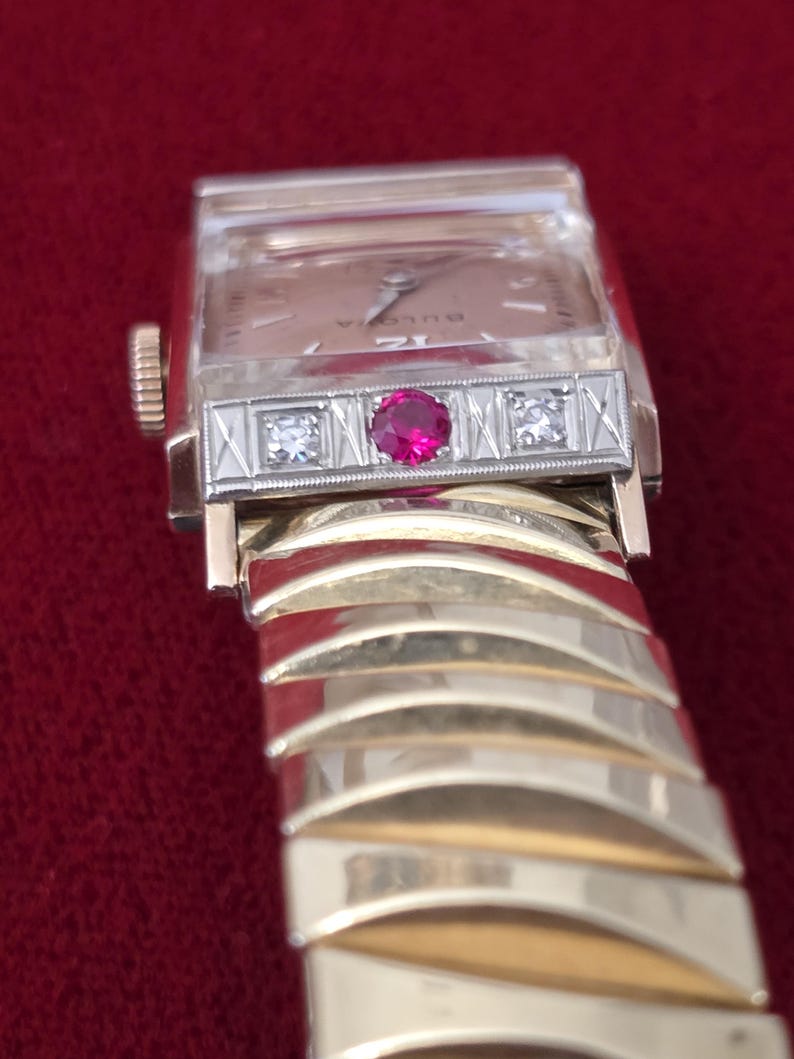 May include: A vintage Bulova wristwatch with a gold-tone band and case. The watch face is rectangular with a light-colored dial. The bezel features a central ruby and two small diamonds. The band is a series of curved gold-tone links.