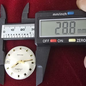Puede incluir: Una esfera de reloj con manecillas y marcadores dorados, etiquetada como "Tradition" y "electronic". Un calibrador digital mide la esfera del reloj a 2,88 cm. El fondo es una tela roja oscura.