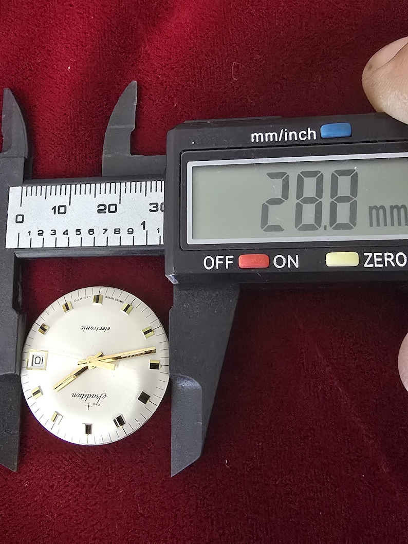 Puede incluir: Una esfera de reloj con manecillas y marcadores dorados, etiquetada como "Tradition" y "electronic". Un calibrador digital mide la esfera del reloj a 2,88 cm. El fondo es una tela roja oscura.