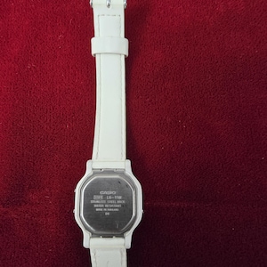 May include: A white Casio LA-11W digital watch with a white leather strap. The watch has a stainless steel back and is water resistant. The watch is displayed against a red background.