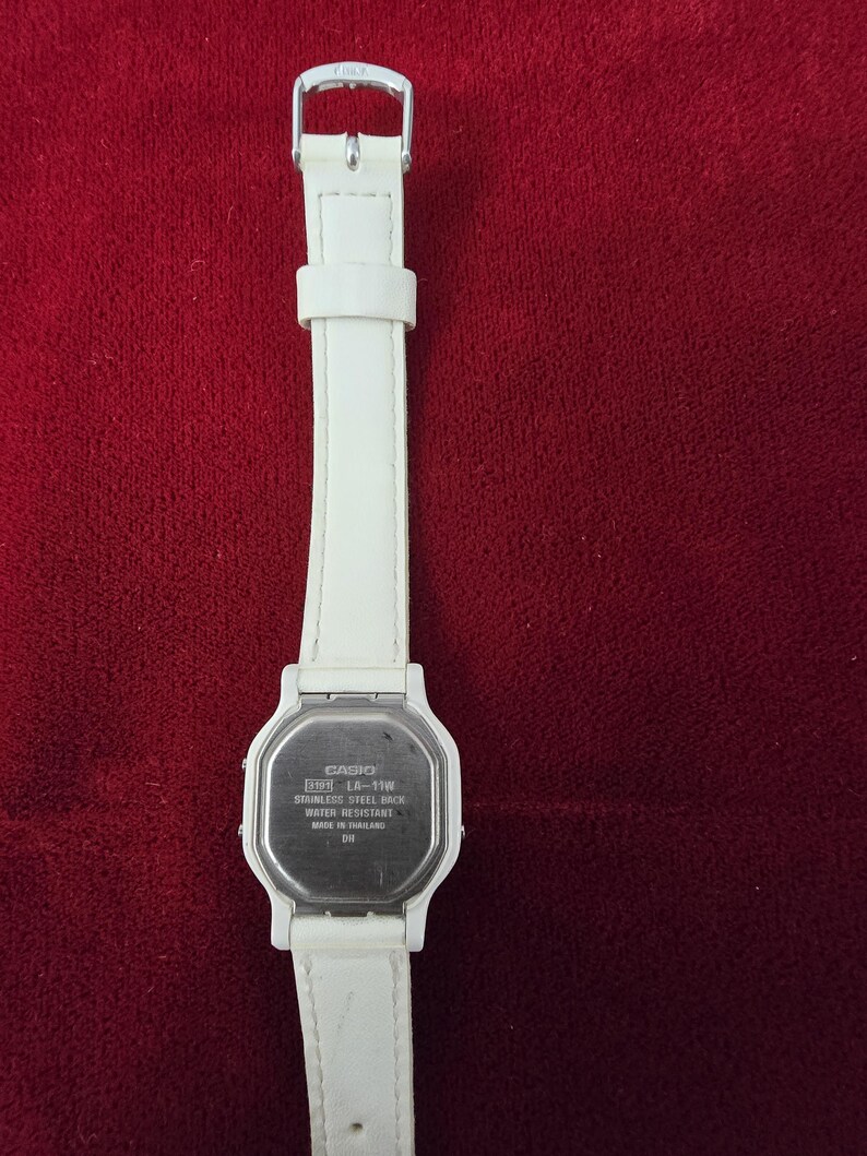 May include: A white Casio LA-11W digital watch with a white leather strap. The watch has a stainless steel back and is water resistant. The watch is displayed against a red background.
