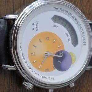 Op de afbeelding: Een zilverkleurig polshorloge met een zwarte leren band met witte stiksels. De witte wijzerplaat toont de tekst THE THIRD MILLENNIUM CHALLENGE, een gele analoge wijzerplaat met Romeinse cijfers, een digitaal display en een paarse bol.