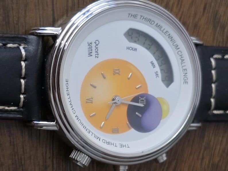 Op de afbeelding: Een zilverkleurig polshorloge met een zwarte leren band met witte stiksels. De witte wijzerplaat toont de tekst THE THIRD MILLENNIUM CHALLENGE, een gele analoge wijzerplaat met Romeinse cijfers, een digitaal display en een paarse bol.