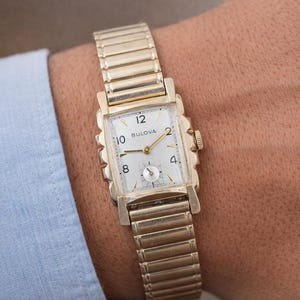 May include: A gold-toned Bulova wristwatch with a rectangular face and a stretch band. The watch face is white with black numerals and gold hands. The watch is on a wrist, with a light blue shirt sleeve visible.