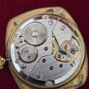 May include: Close-up of a vintage gold-toned watch with the back removed, revealing the intricate mechanical movement. The watch mechanism includes gears, jewels, and the text "EBEL 107" and "17 JEWELS SWISS UNADJUSTED".