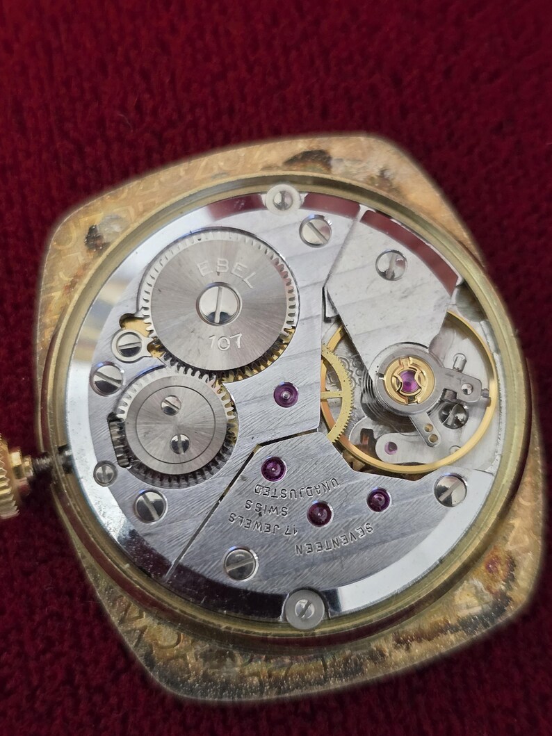 May include: Close-up of a vintage gold-toned watch with the back removed, revealing the intricate mechanical movement. The watch mechanism includes gears, jewels, and the text "EBEL 107" and "17 JEWELS SWISS UNADJUSTED".