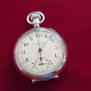 May include: A silver pocket watch with a white face and black numerals. The watch has a small seconds dial and the words "NEW YORK STANDARD WATCH CO. U.S.A." are printed on the face. The watch is set against a red background.