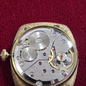 May include: A vintage gold-toned watch with the back removed, revealing the intricate mechanical movement. The watch mechanism features gears, jewels, and the inscription "7 JEWELS". The watch has a blue gemstone on the winding stem.