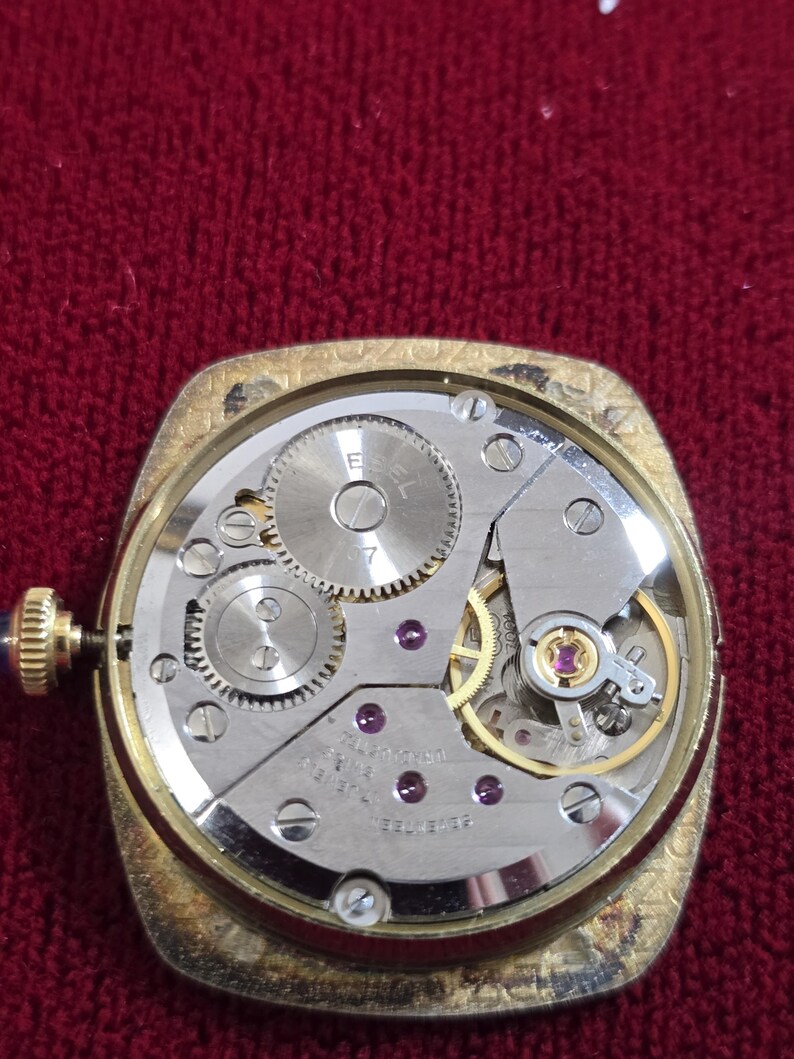 May include: A vintage gold-toned watch with the back removed, revealing the intricate mechanical movement. The watch mechanism features gears, jewels, and the inscription "7 JEWELS". The watch has a blue gemstone on the winding stem.
