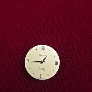K&ouml;nnte beinhalten: Ein goldfarbenes Zifferblatt mit schwarzen Zeigern und r&ouml;mischen Ziffern. Auf dem Zifferblatt stehen die Markennamen "OMEGA" und "DE VILLE QUARTZ". Ein kleiner Diamant ist auf der 12-Uhr-Position gesetzt.
