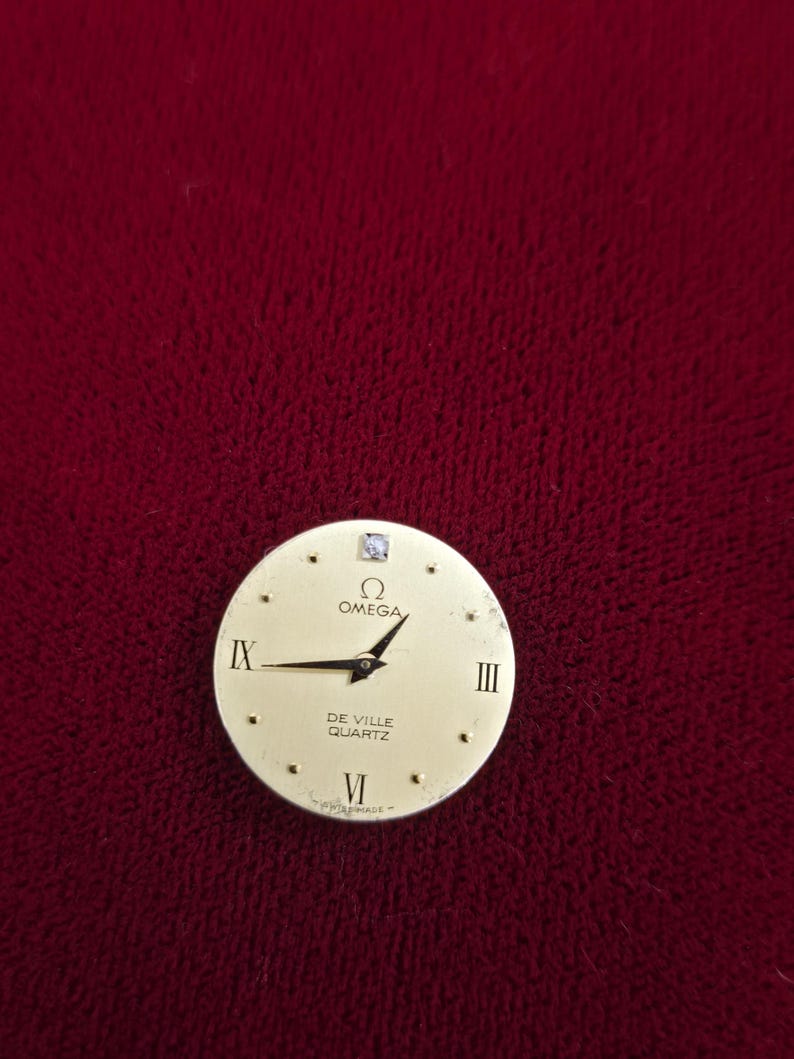 K&ouml;nnte beinhalten: Ein goldfarbenes Zifferblatt mit schwarzen Zeigern und r&ouml;mischen Ziffern. Auf dem Zifferblatt stehen die Markennamen "OMEGA" und "DE VILLE QUARTZ". Ein kleiner Diamant ist auf der 12-Uhr-Position gesetzt.