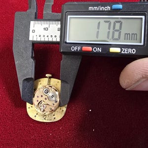 Pu&ograve; includere: Un movimento di orologio dorato, di circa 1,78 cm di diametro, viene misurato con un calibro digitale. Il meccanismo dell'orologio presenta ingranaggi intricati e rubini. Il calibro mostra la misura in millimetri.